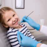 Blonde girl in a striped shirt smiles in the dentist chair while her dentist checks her teeth
