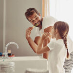Dad in white t-shirt smiles as he brushes his teeth next to his daughter at the bathroom sink